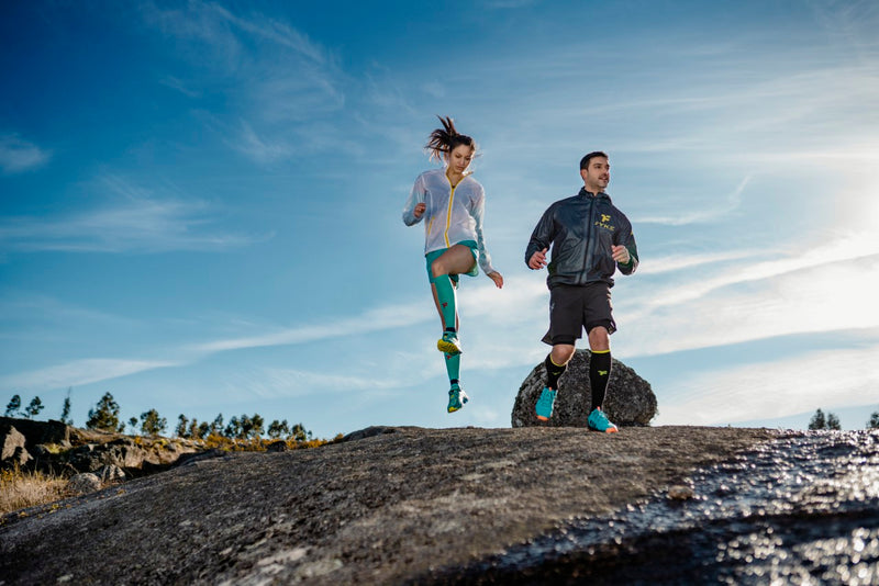 Un homme court et une femme saute dans une zone rocheuse en portant des vêtements de sport Fyke.