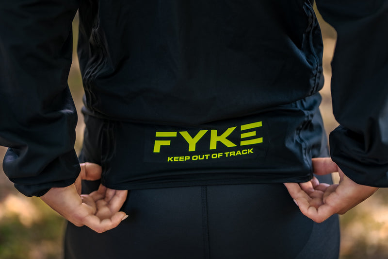 Une femme étire le dos de l'une des vestes de pluie de course à pied de Fyke, montrant le logo de la marque.