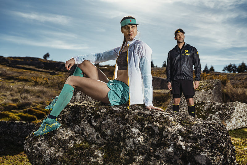 Mulher sentada numa rocha e homem de pé, numa paisagem montanhosa, com roupa desportiva Fyke.